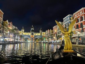 Golden statue with wings on a boat on a canal, illuminated colorful buildings along both banks at night with arches in the distance and reflections on the water.