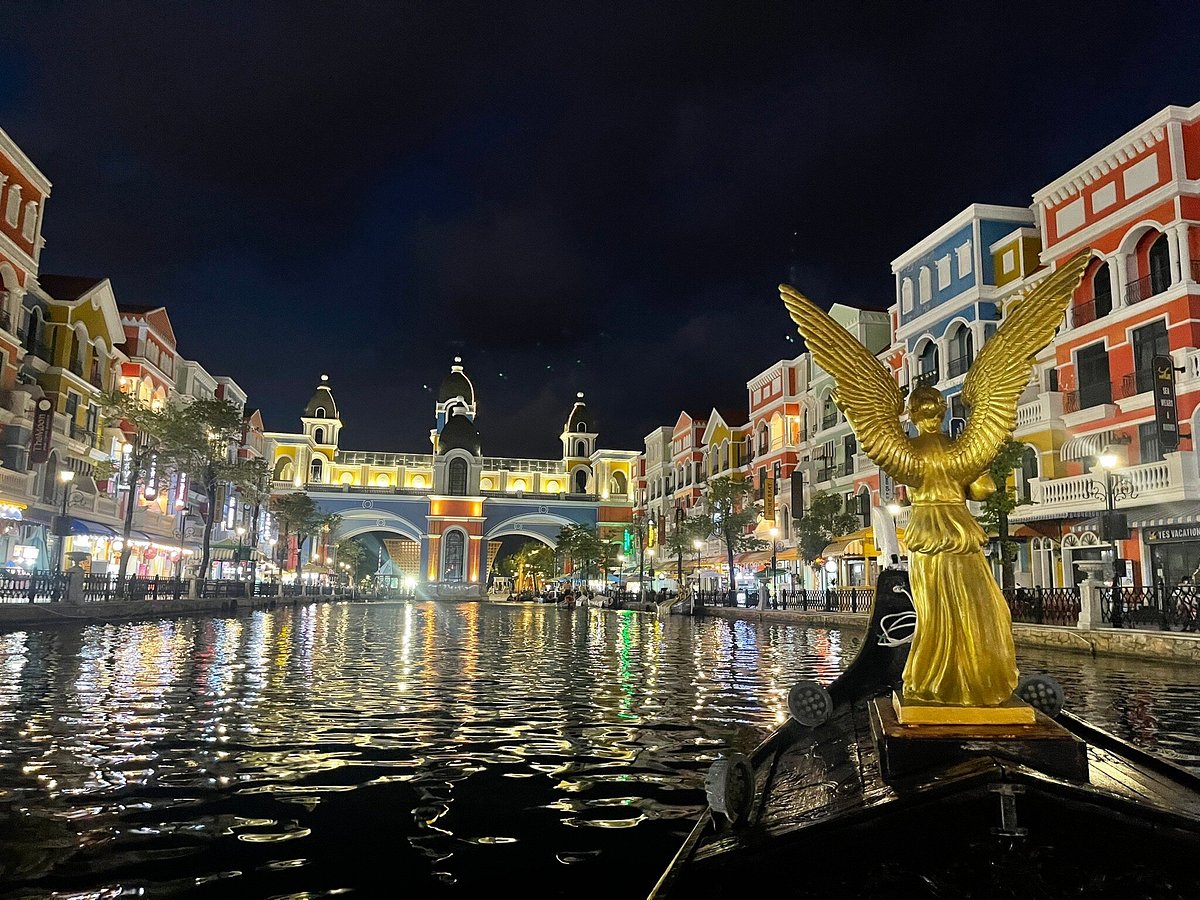 Golden statue with wings on a boat on a canal, illuminated colorful buildings along both banks at night with arches in the distance and reflections on the water.