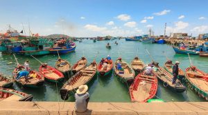 Row of wooden fishing boats tied to a concrete dock in a sunny harbor, fishermen at work on calm turquoise water.