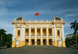 Grand neoclassical building with yellow and white facade, tall columns, and steps leading to arched entrances; Vietnamese flag flies atop the roof.