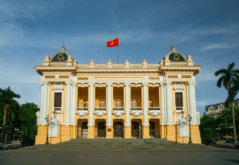 Grand neoclassical building with yellow and white facade, tall columns, and steps leading to arched entrances; Vietnamese flag flies atop the roof.