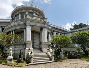 White neoclassical building with a circular porte-cochère, grand steps, and columns at the entrance; garden with lion statues nearby.