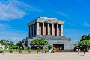Front view of Ho Chi Minh Mausoleum with tall stone columns, a clear blue sky, and trimmed shrubs in a wide plaza; visitors near the entrance.