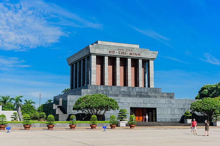 Front view of Ho Chi Minh Mausoleum with tall stone columns, a clear blue sky, and trimmed shrubs in a wide plaza; visitors near the entrance.