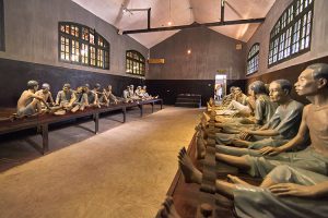 Life-size statues of people seated along long dining tables in a dim museum hall, facing each other as if sharing a meal.