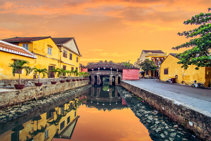 Golden sunset over a canal in a historic town, with yellow buildings, palm planters, and a decorative arched bridge reflecting in the water.