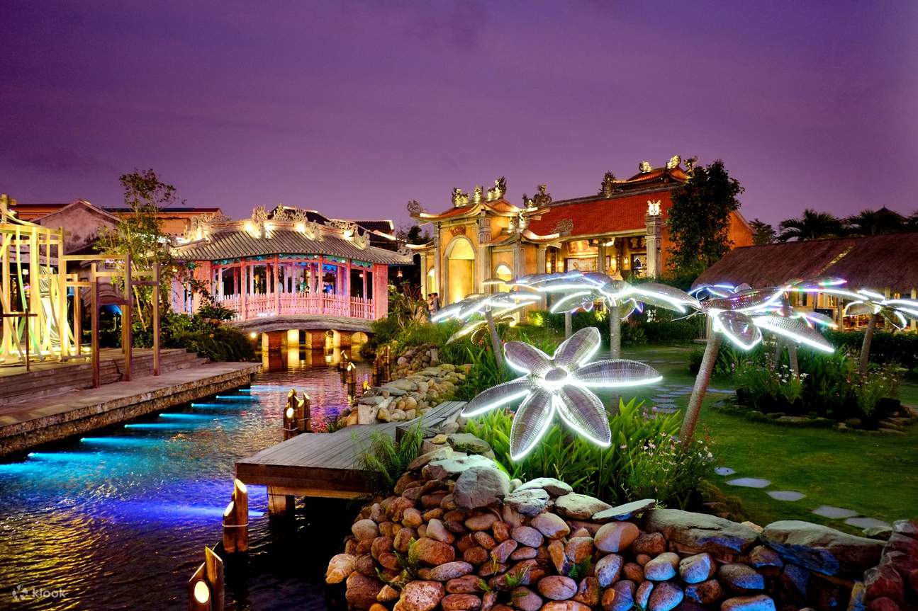 Night view of a landscaped garden with glowing flower light sculptures, a canal, and traditional buildings illuminated in warm colors.