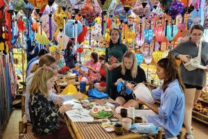 Group of people around a wooden table sewing and painting paper lanterns in a shop decorated with hundreds of colorful lanterns overhead.