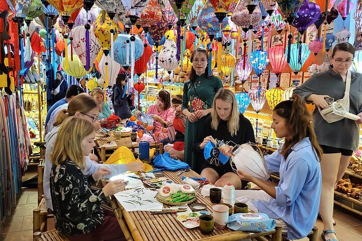 Group of people around a wooden table sewing and painting paper lanterns in a shop decorated with hundreds of colorful lanterns overhead.