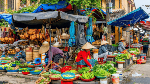 Vibrant street market with vendors wearing conical hats selling greens and vegetables in baskets under colorful awnings.