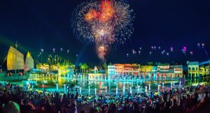 Nighttime festival with colorful fireworks above a water stage and performers in white costumes, with a seated audience around the basin.