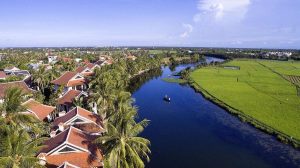 Aerial view of a tropical village with red-roofed houses along a winding river, palm trees, and green fields under a blue sky.