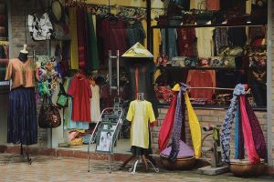 Colorful clothing storefront with mannequins wearing bright dresses and a yellow tunic, plus racks of fabrics visible in the window.