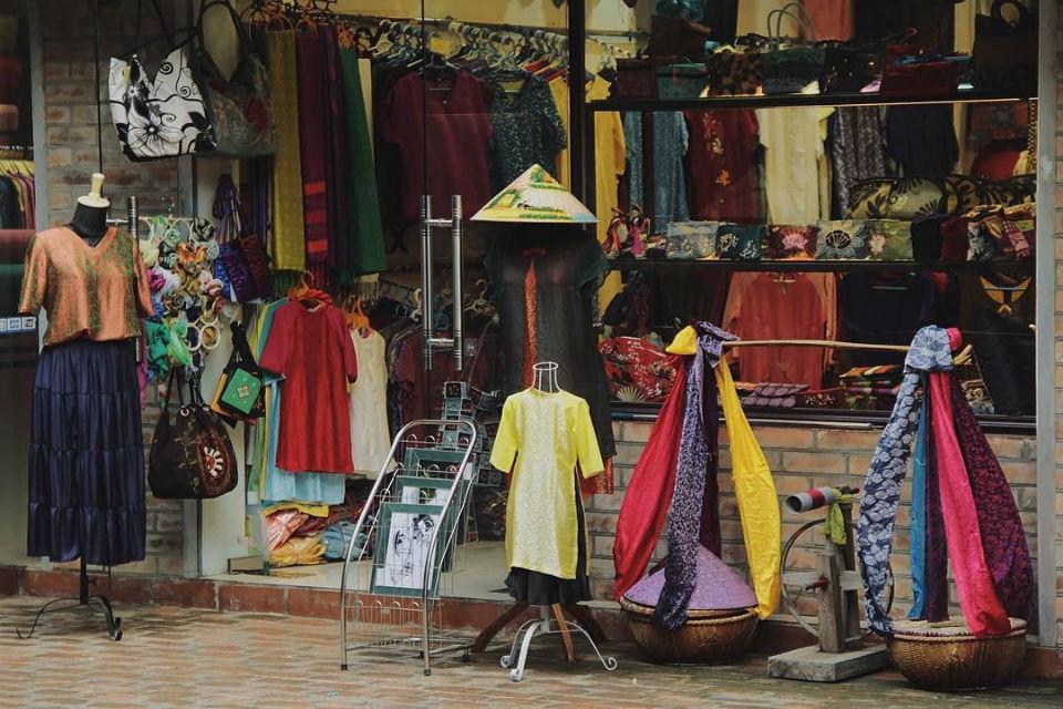 Colorful clothing storefront with mannequins wearing bright dresses and a yellow tunic, plus racks of fabrics visible in the window.