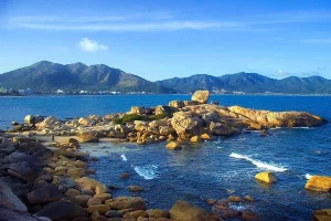 Rocky coastline extending into calm blue sea with mountains in the distance under a clear sky.