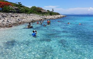 Clear turquoise water along a rocky beach with people swimming; a person in blue holds a camera in the foreground, while thatched huts line the shore.