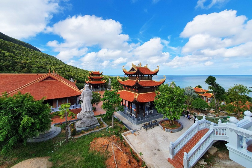 Coastal Chinese temple complex with red tiered pagodas, a white statue, and a white bridge overlooking the sea.