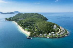 A tropical island resort on a green peninsular with a white-sand beach, palm trees, and rows of villas along a curved shoreline beside turquoise water.