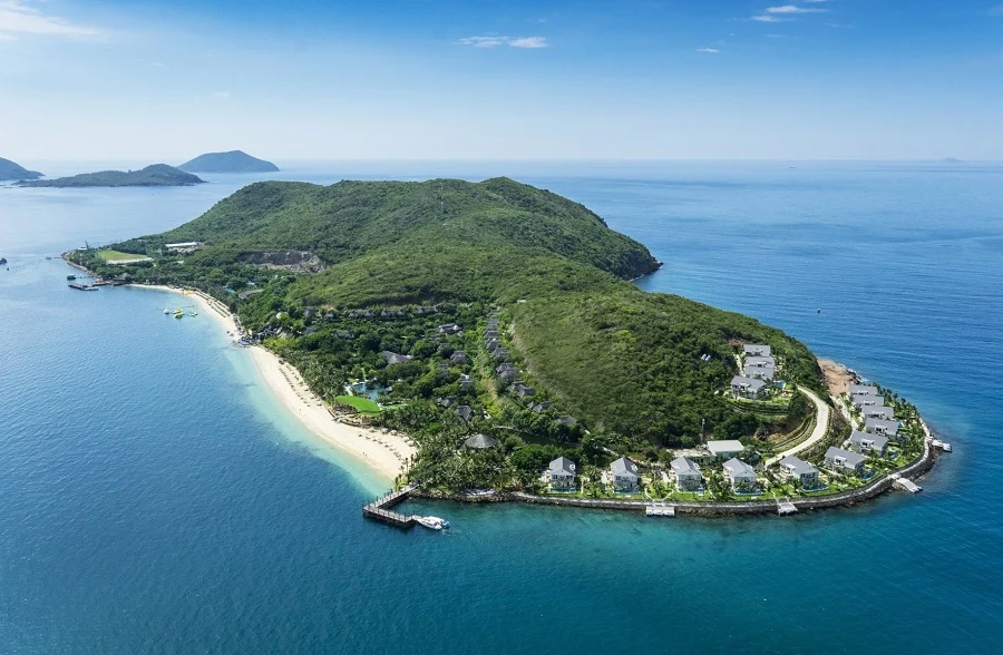 A tropical island resort on a green peninsular with a white-sand beach, palm trees, and rows of villas along a curved shoreline beside turquoise water.