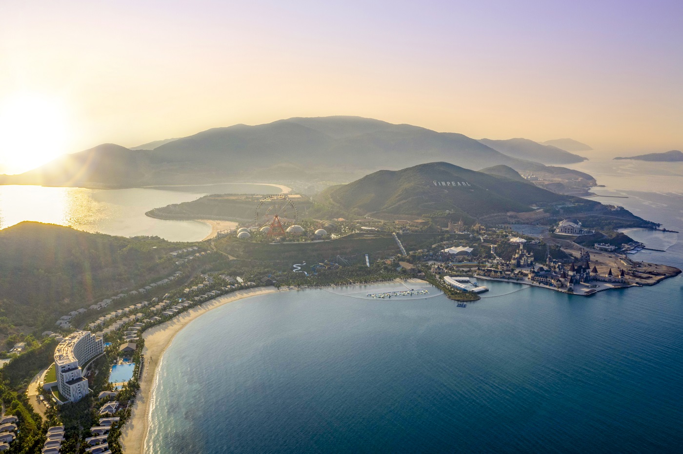 Aerial view of a coastal resort at sunset, with a curved sandy beach, blue bay, and distant hills. A ferris wheel sits near the center.
