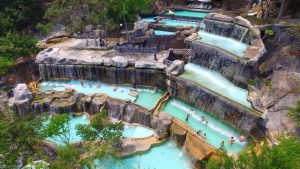 A multi-tiered turquoise water park with cascading waterfalls and people swimming in the pools.