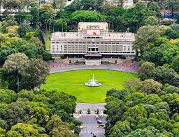 Aerial view of a large neoclassical building surrounded by a green park, with a central circular fountain on a manicured lawn.