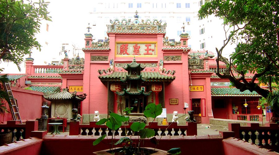 Red Chinese temple with green tiled roofs and a central ornate entrance, flanked by guardian lions in a courtyard with lotus plants in the foreground.
