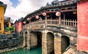 Stone arched bridge over a turquoise canal, with a wooden walkway and red lanterns in a colorful riverside town.