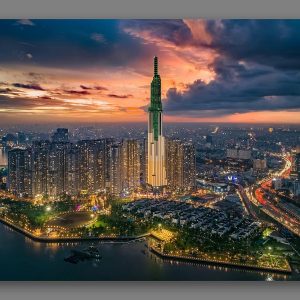 Aerial view of a green-lit skyscraper towering over a dense city at sunset with a riverfront park in the foreground.