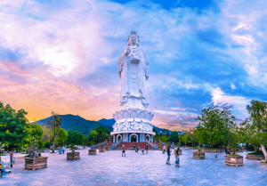 Large white statue of Guanyin atop a lotus pedestal in a spacious plaza at dusk, with visitors and trees around and mountains in the background.