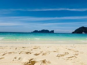 Sunny tropical beach with pale sand, turquoise water, and distant rocky islets under a clear blue sky.