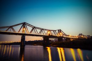 Evening view of a steel truss bridge lit with orange lights spanning a calm river, reflections glowing on the water.