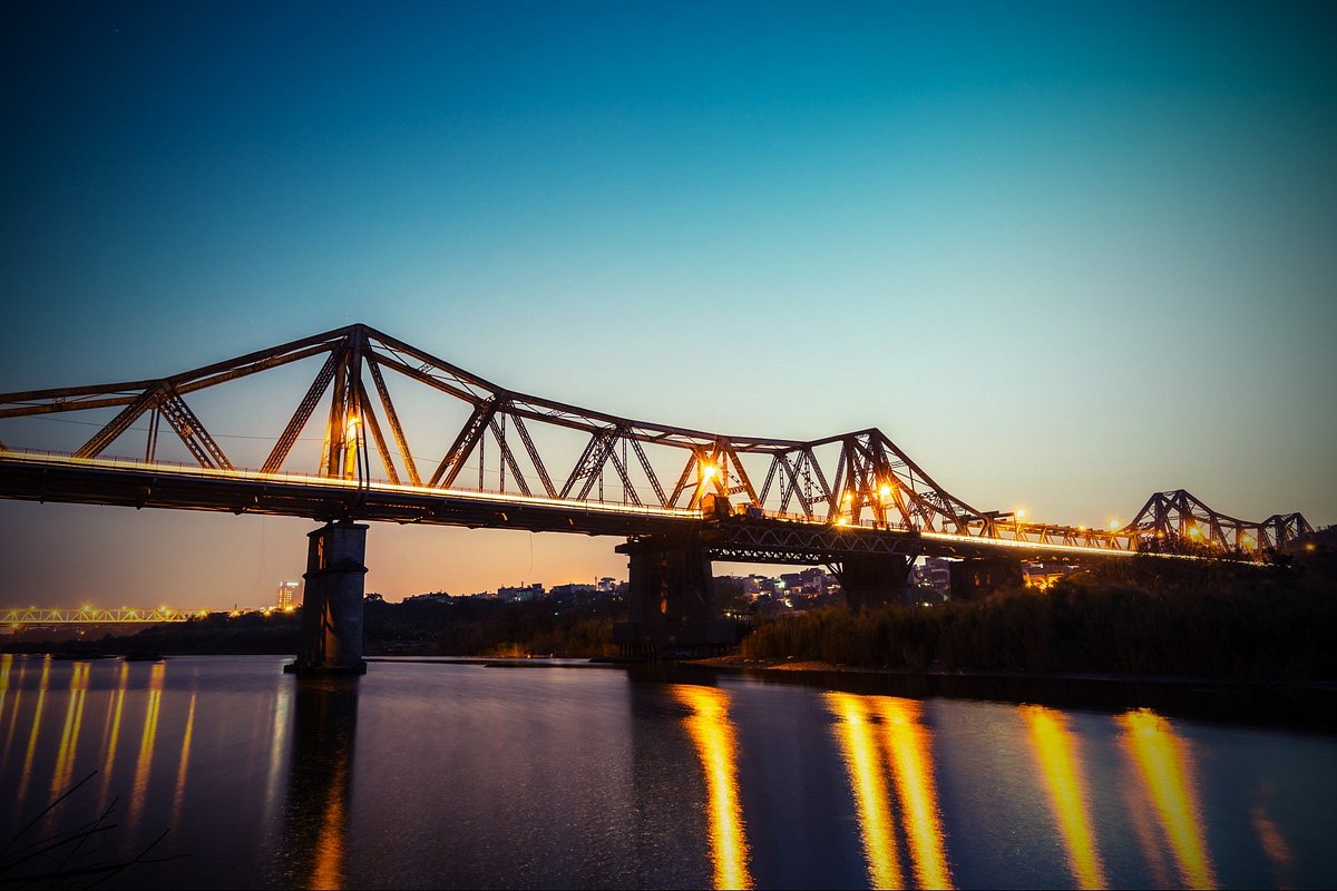 Evening view of a steel truss bridge lit with orange lights spanning a calm river, reflections glowing on the water.
