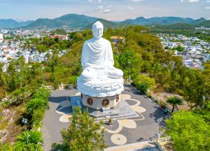 Large white Buddha statue seated in meditation on a circular tiled platform, with a city and distant mountains behind.