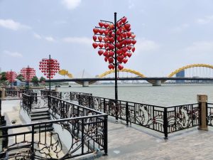 Riverside walkway with black decorative railing and red heart lanterns on poles, yellow bridge in distance and a clear sky.