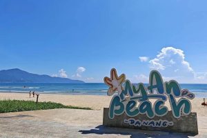 My An Beach sign in Da Nang on a sunny beach with blue sea and distant mountains in the background, sign lettering colorful and playful