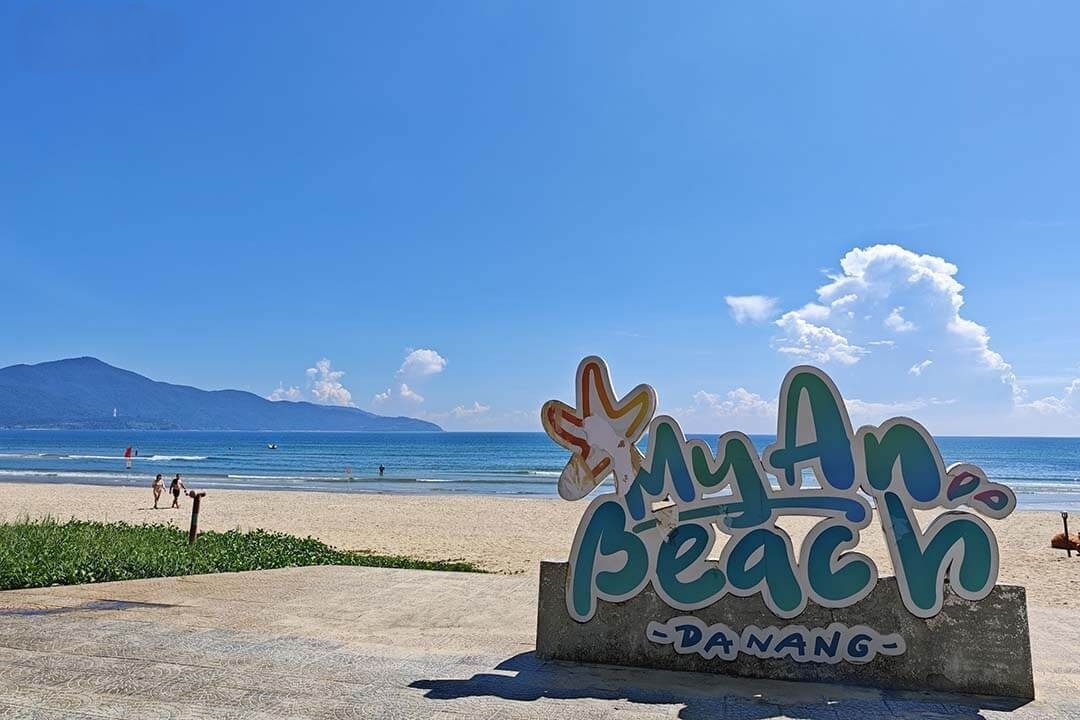 My An Beach sign in Da Nang on a sunny beach with blue sea and distant mountains in the background, sign lettering colorful and playful