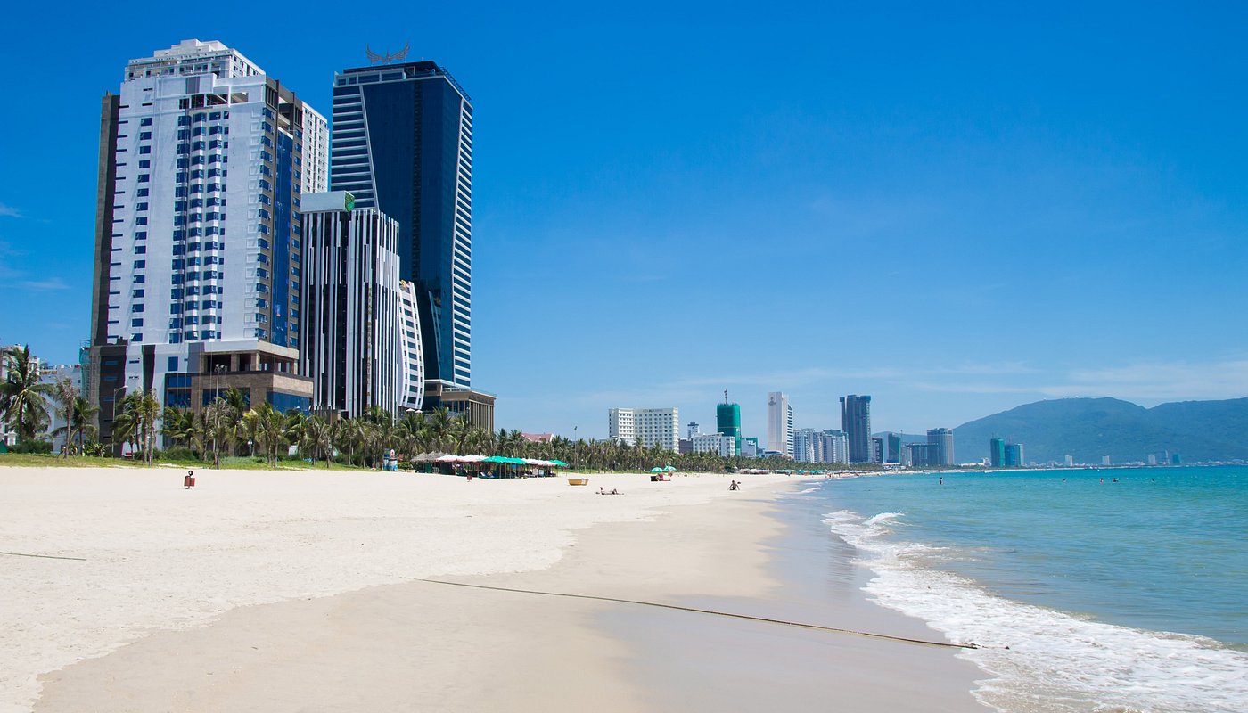 Wide city beach with white sand and turquoise water, tall modern buildings along the shore under a clear blue sky.