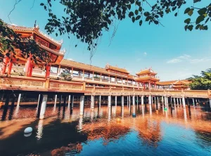 Traditional Chinese-style temple complex with ornate red buildings on a wooden walkway over calm water, reflected in the lake.