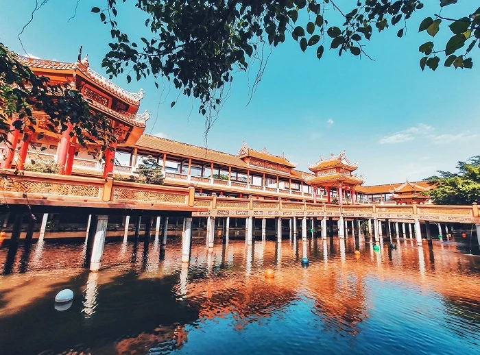 Traditional Chinese-style temple complex with ornate red buildings on a wooden walkway over calm water, reflected in the lake.