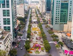 Aerial view of a city boulevard with modern high-rise buildings on both sides, featuring a tree-lined median and circular flower beds at the center.