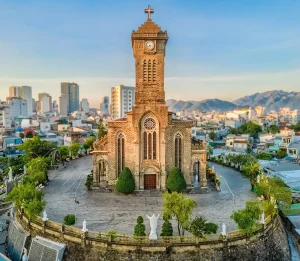 Stone church with tall clock tower and cross atop, set on a hill overlooking a city and distant mountains.