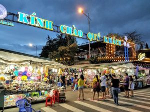 Night market at dusk with stalls selling hats, colorful goods, and string lights under a large Vietnamese sign welcoming guests: 'Kinh chao quy khach'