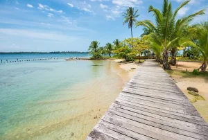 Wooden boardwalk along a tropical beach with palm trees, turquoise water, and a small boat anchored near shore.