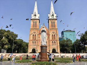 A red-brick church with two tall white spires and a statue in front, in a busy city square with pigeons in the sky.