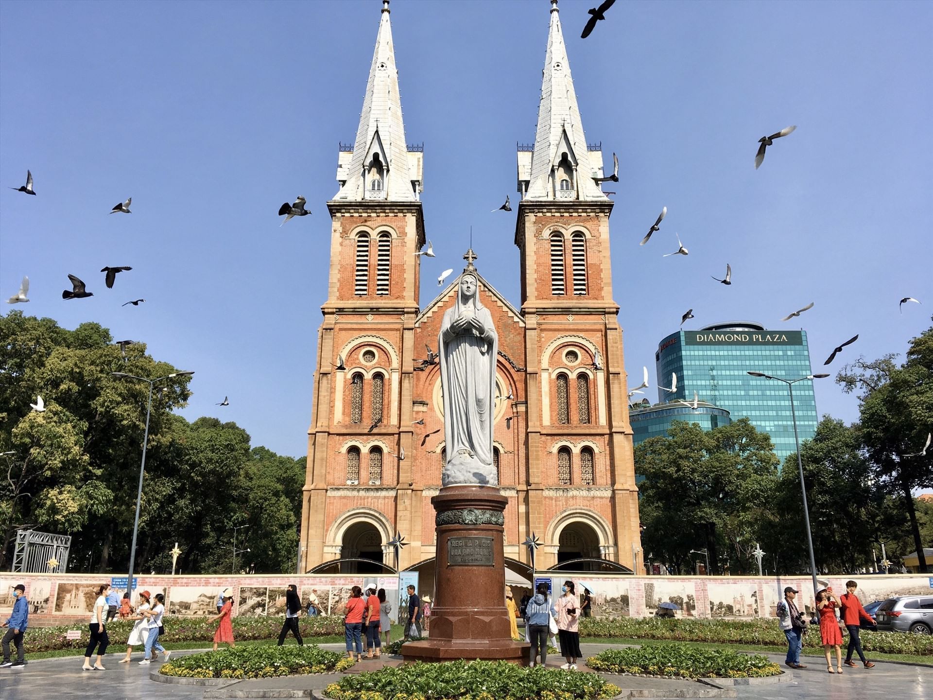 A red-brick church with two tall white spires and a statue in front, in a busy city square with pigeons in the sky.