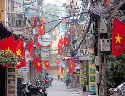 Narrow Vietnamese street lined with red flags featuring yellow stars, storefronts on both sides, motorcycles parked along the road, and tangled overhead wires.
