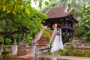 Traditional wooden pavilion on stilts over a pond, with a curved tiled roof and stone steps surrounded by green trees.