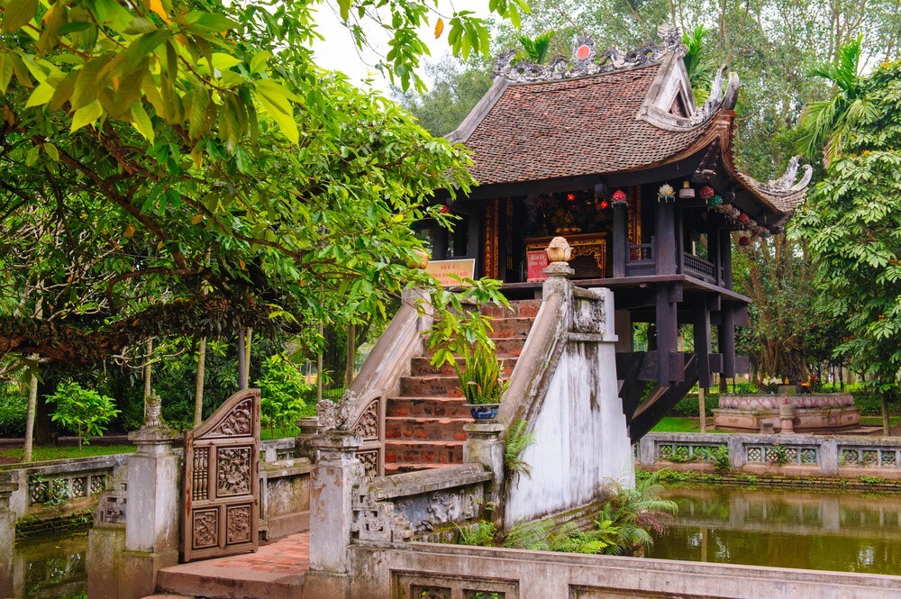 Traditional wooden pavilion on stilts over a pond, with a curved tiled roof and stone steps surrounded by green trees.