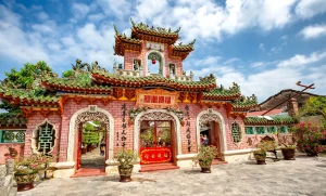 Colorful Chinese temple entrance with red walls, green tiled roofs, and ornate carvings under a blue sky with clouds.